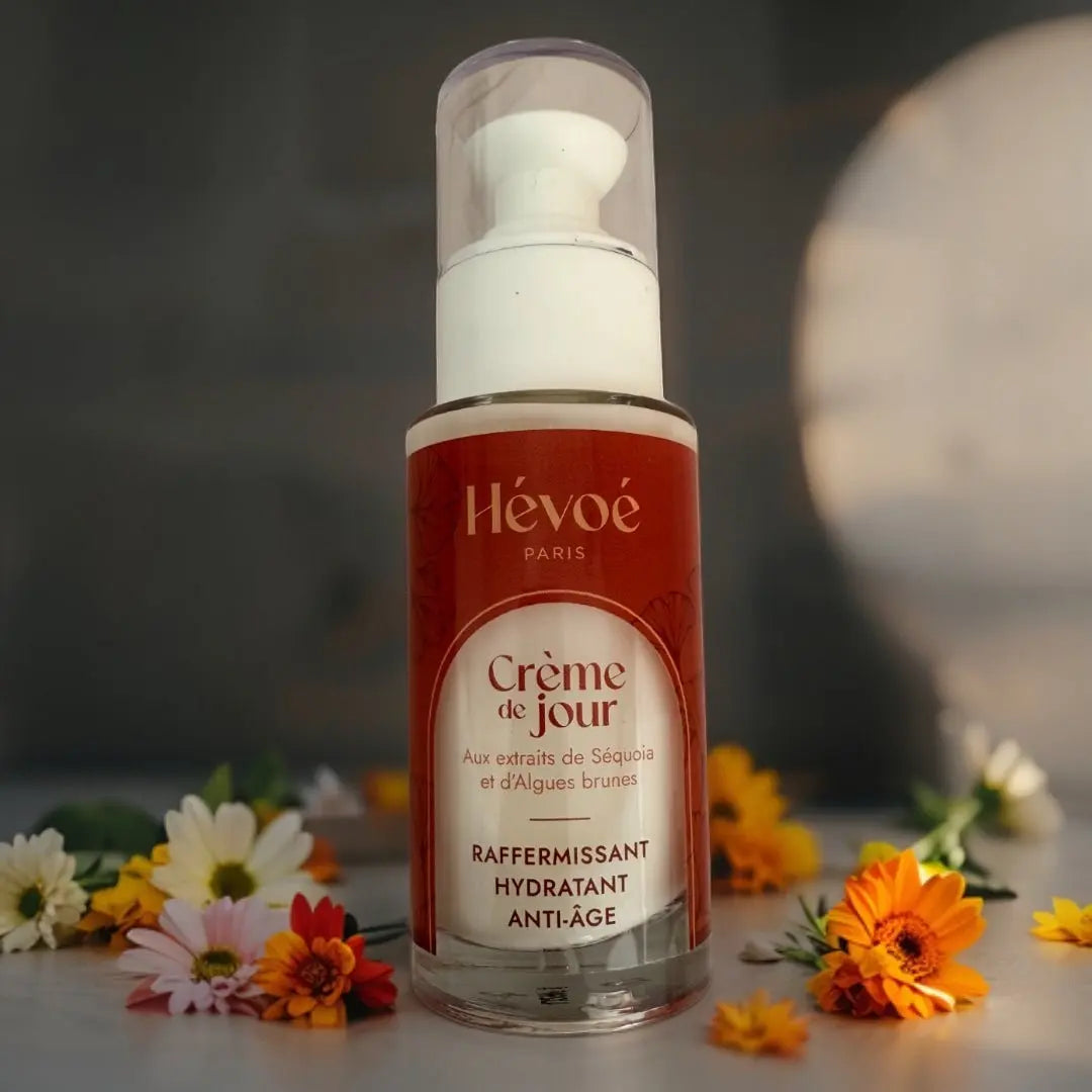 HEVOE Crème de Jour Raffermissant Hydratant Anti-Âge, flacon pompe en verre avec étiquette rouge et dorée, entouré de fleurs colorées.